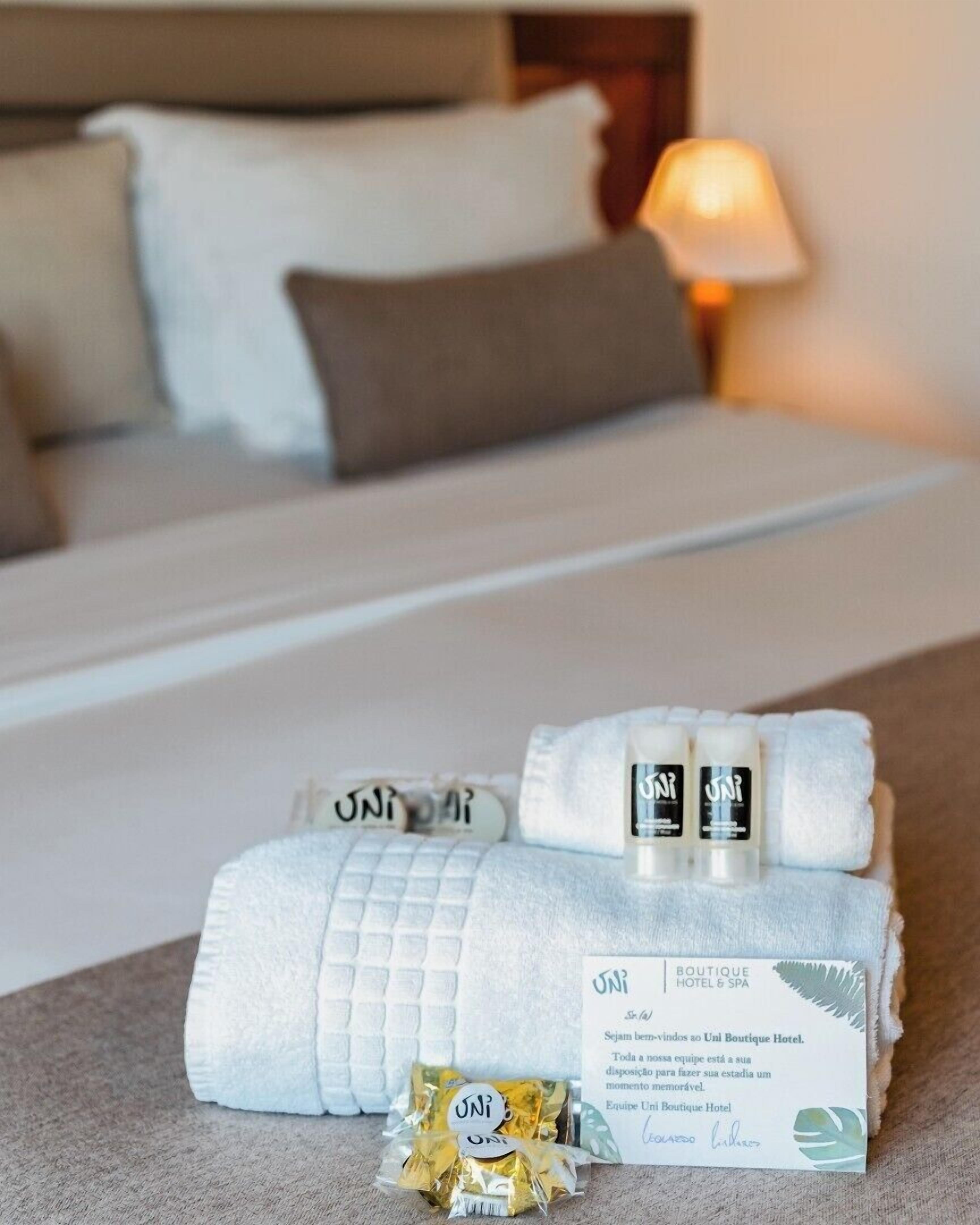 A neatly made bed with a white duvet, a folded towel, and a small package labeled "UNI" on a hotel bed.