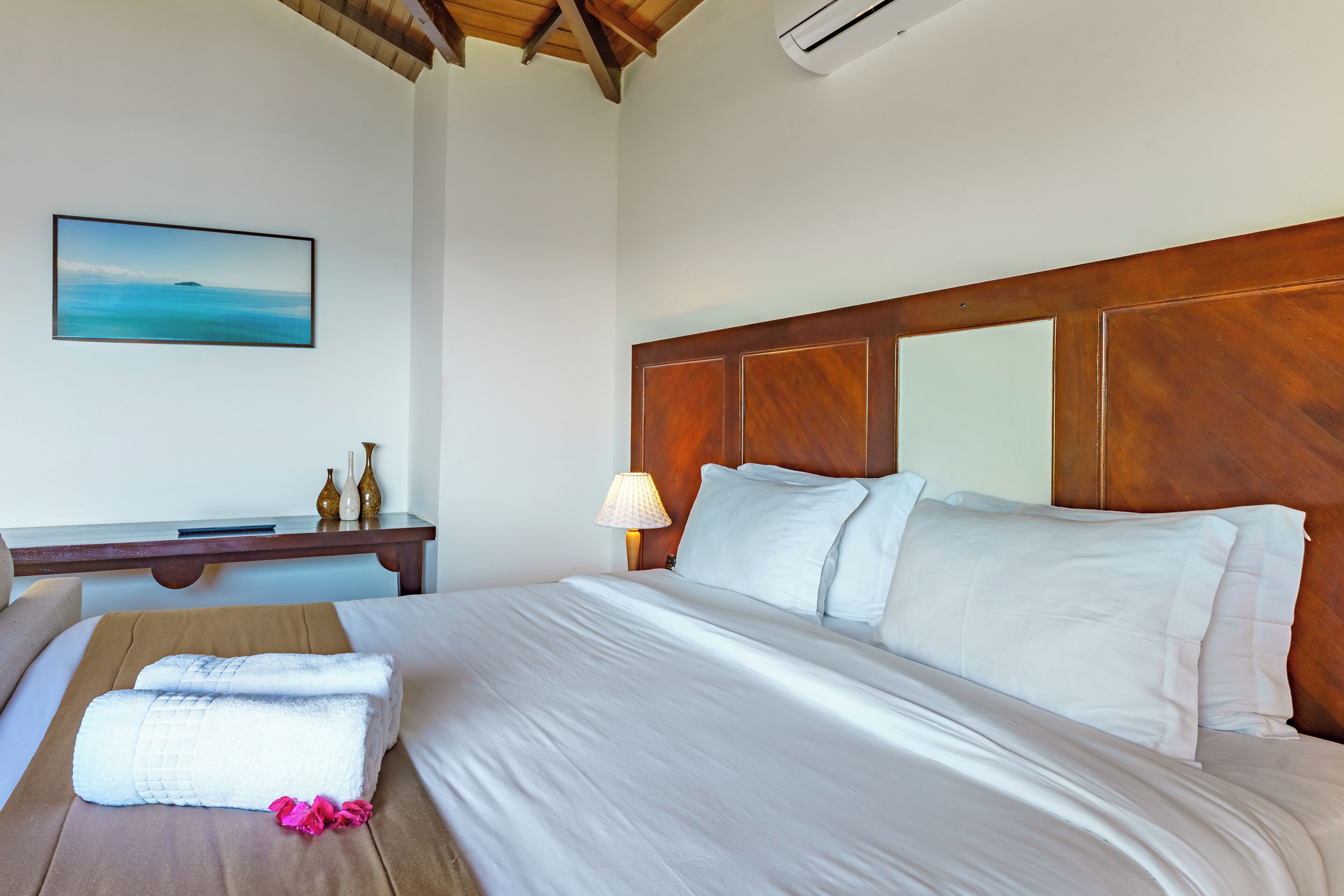 A neatly made bed with white linens and a wooden headboard, a bedside table with a lamp, and a framed picture of a coastal scene on the wall.