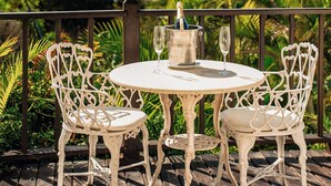 Une table ronde avec deux chaises, placée sur une terrasse en bois, d’où l’on peut admirer un paysage côtier avec des collines et une plage.