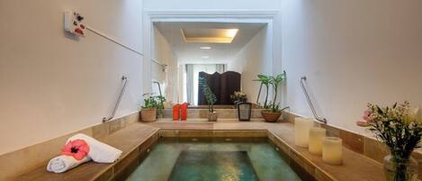 A spa area with a large soaking tub, wooden benches, potted plants, and a view of a room with a curtain.