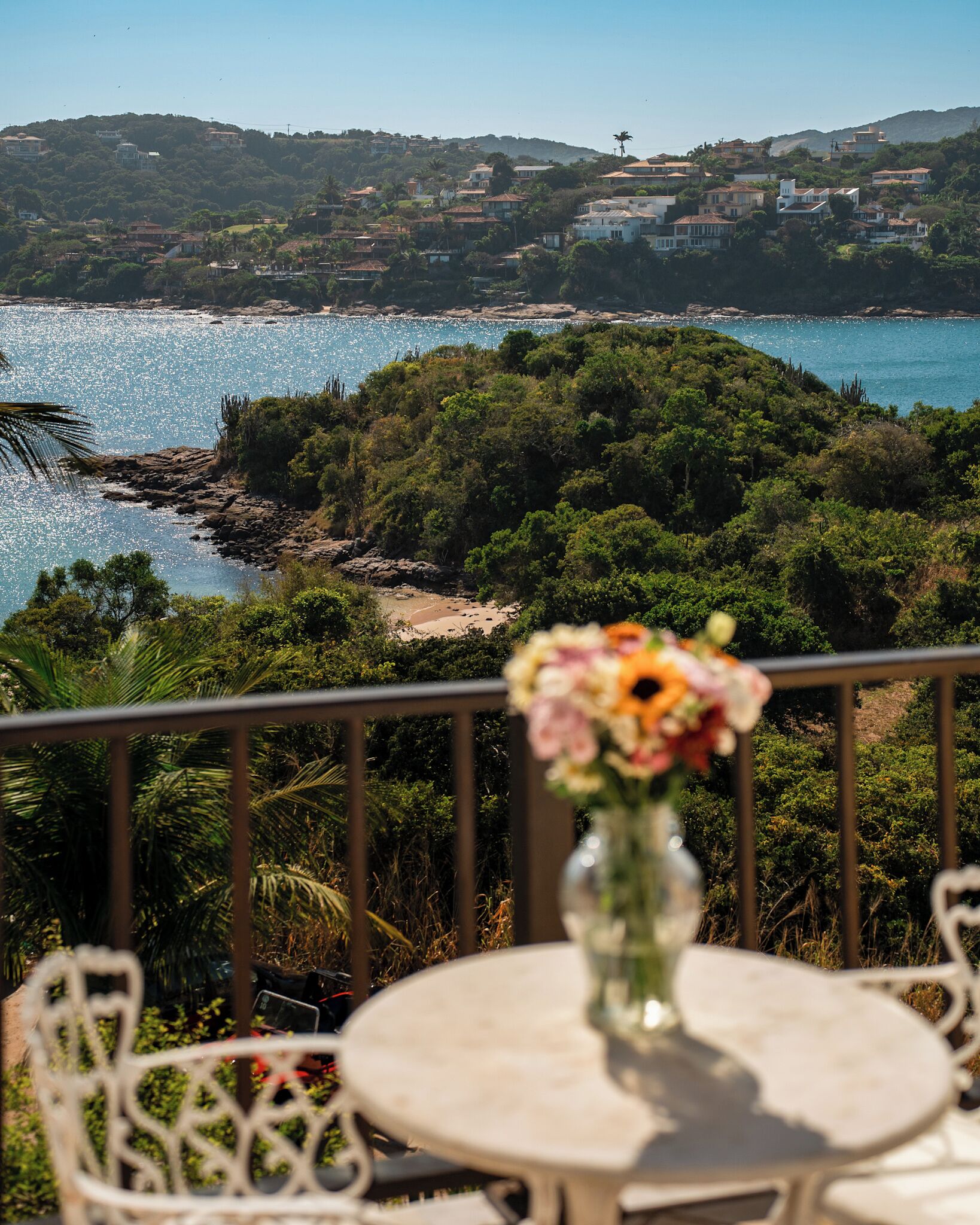 Un balcón con mesa y sillas, con vistas a una zona costera con casas y vegetación.