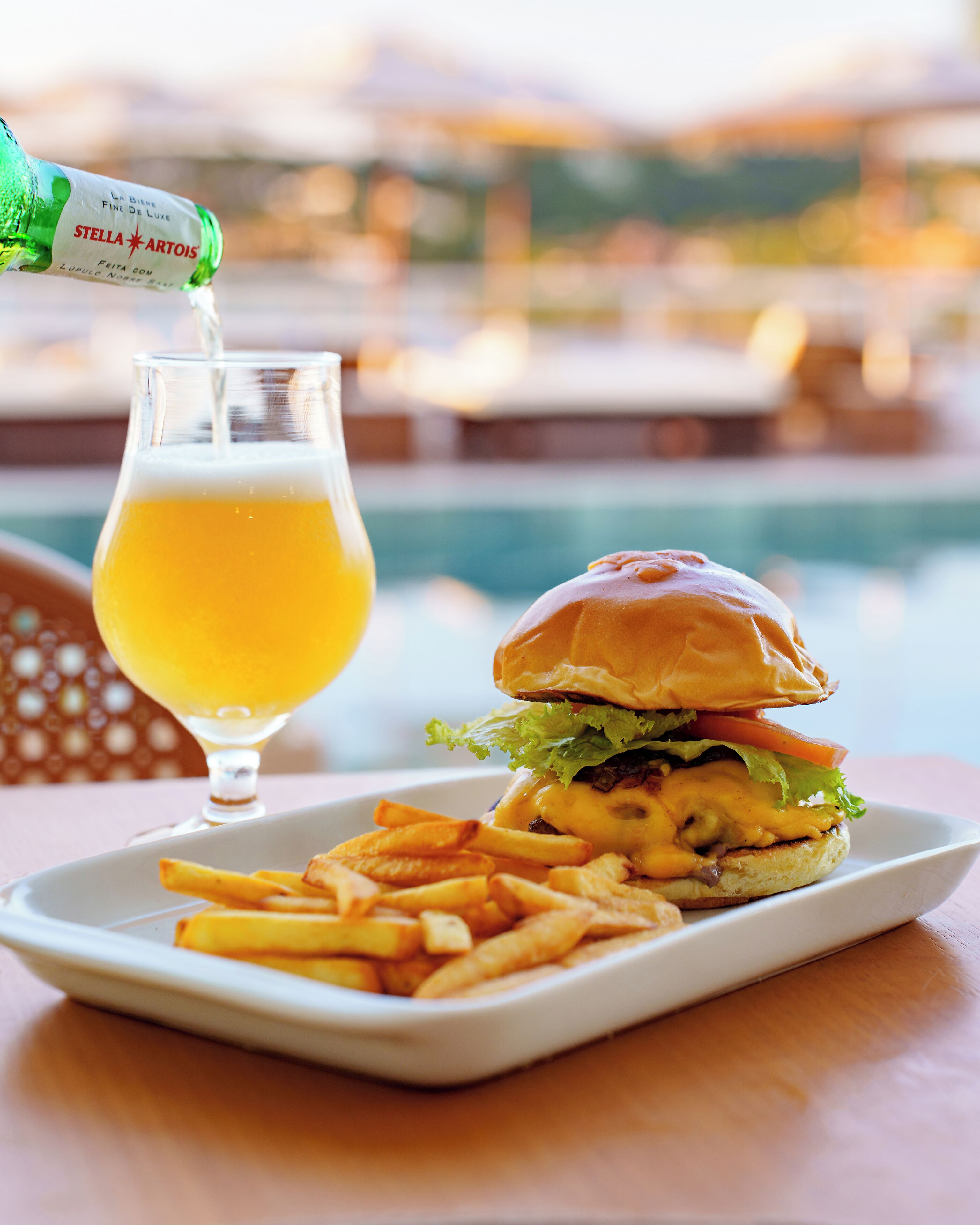 Eine Mahlzeit bestehend aus einem Burger, Pommes und einem Glas Bier, wobei ein Flaschenbier von Stella Artois ins Glas eingeschenkt wird.