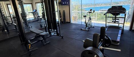 Uma academia com equipamentos de musculação, esteira e vista para o mar.