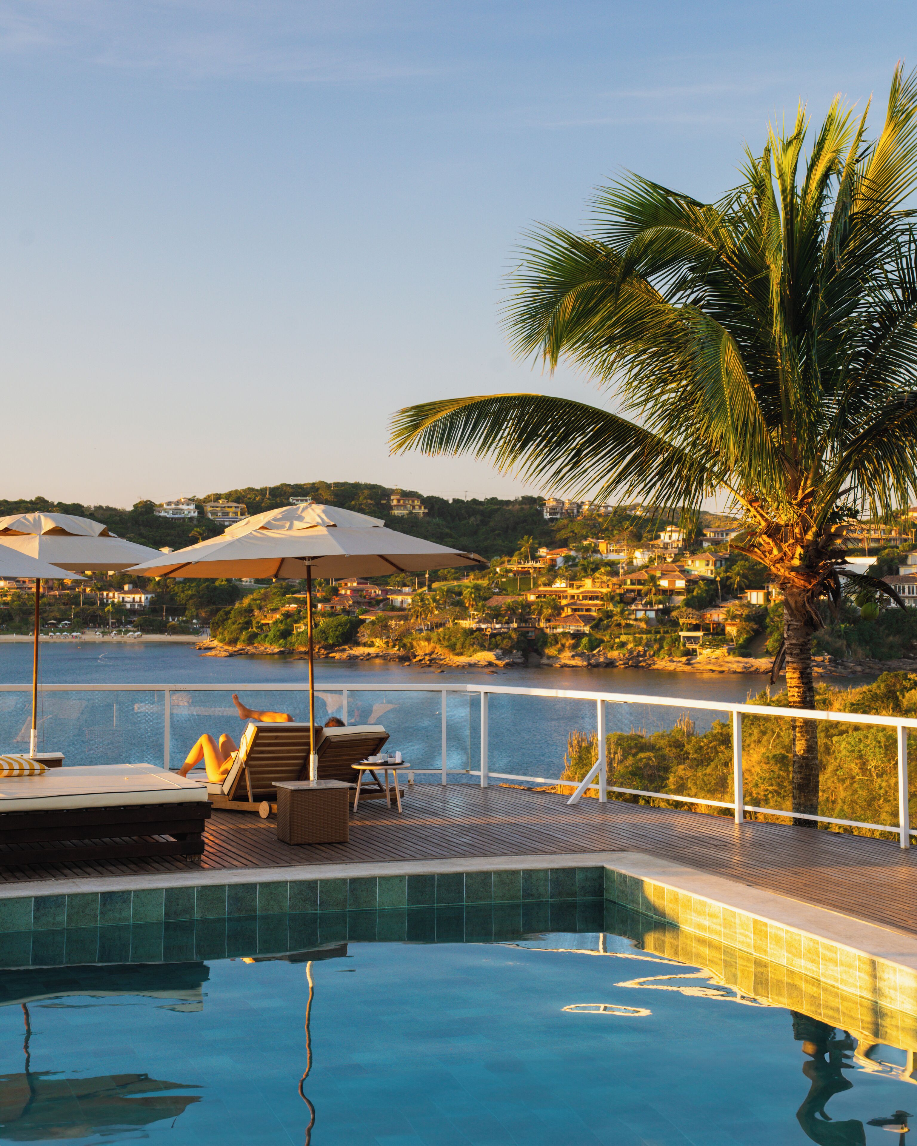 A pool area with lounge chairs and umbrellas, a view of the sea, and a palm tree.