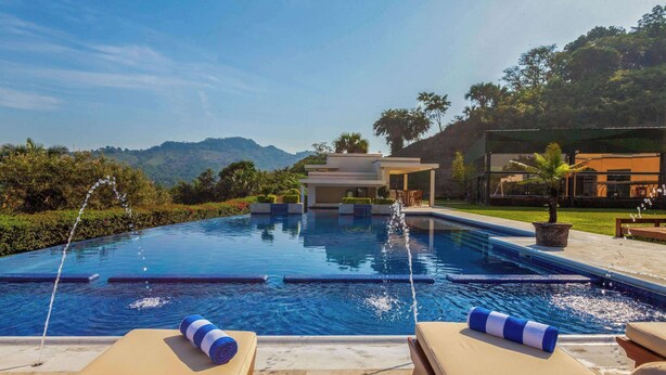 A swimming pool with lounge chairs and a covered area, surrounded by greenery and hills.