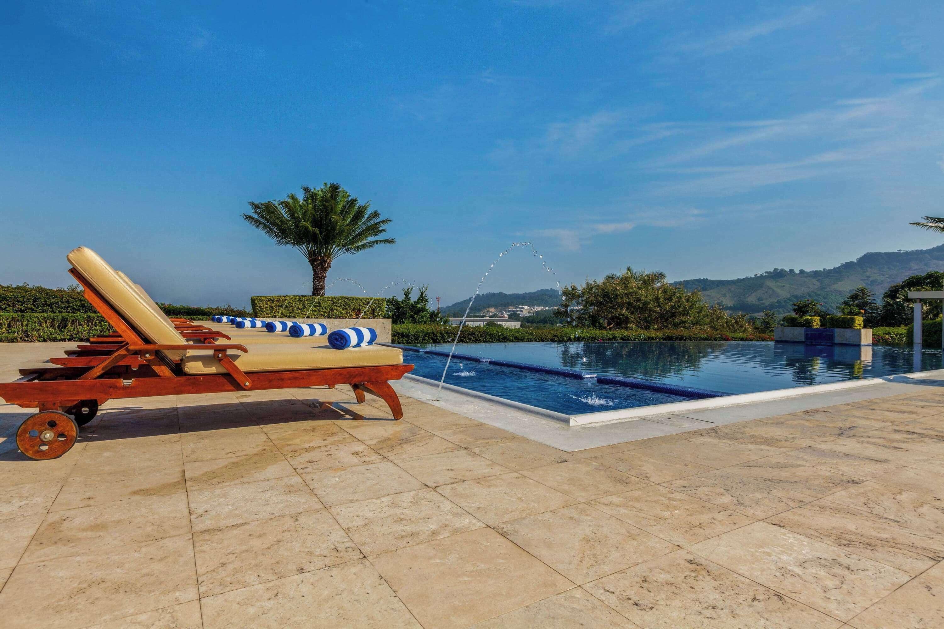 A poolside area with a lounge chair and a small table, surrounded by a landscaped garden and a view of hills.
