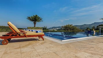Zona junto a la piscina con una silla de descanso y una mesita, rodeada de un jardín ajardinado y vistas a colinas.