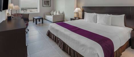 A hotel room with a large bed, a desk, a chair, a sofa, and a TV.