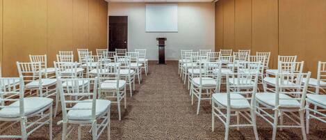 Un salón de conferencias con filas de sillas blancas, piso alfombrado y una pantalla grande al frente.