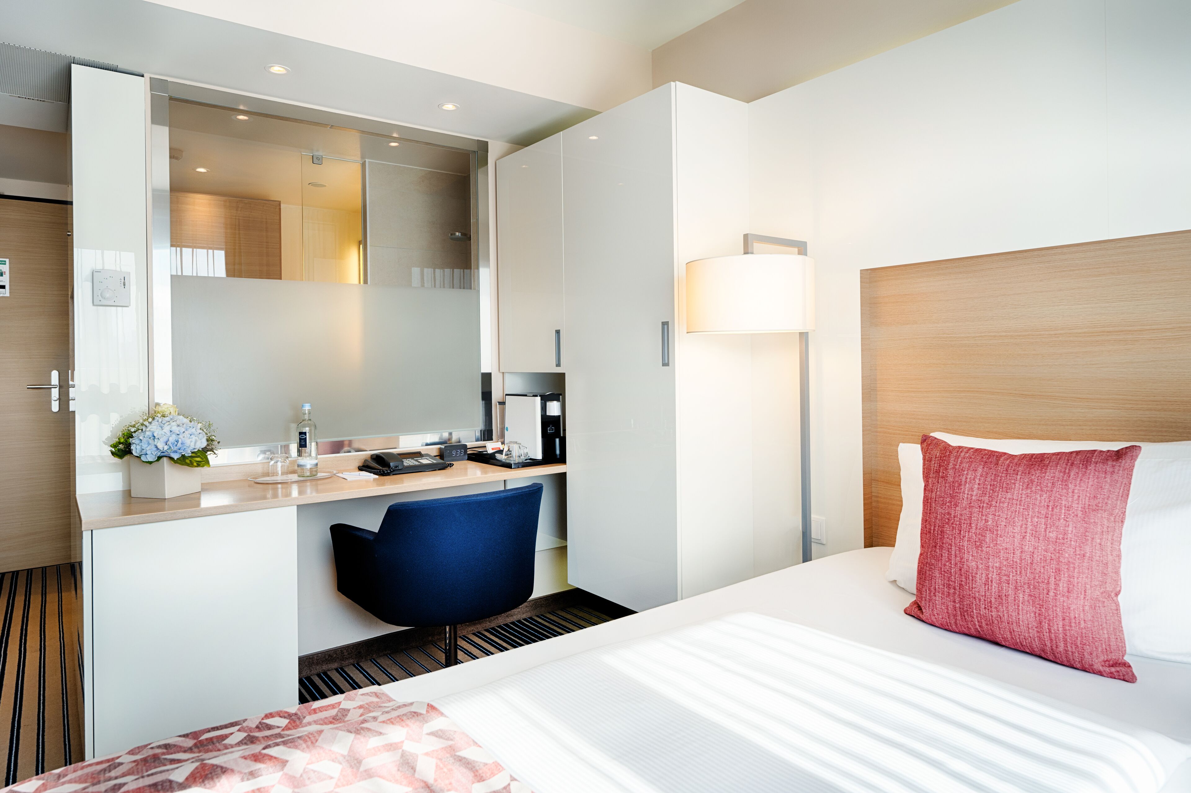 A hotel room with a bed, a desk with a chair, a mirror, a lamp, and a small table with a vase of flowers.