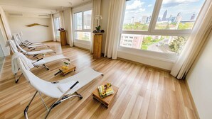 A room with wooden flooring, white chairs, a table with food, and large windows with a city view.