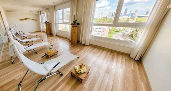 Una habitación con piso de madera, sillas blancas, una mesa con comida y ventanas amplias con vista a la ciudad.