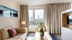 A hotel room with a sofa, a coffee table, a vase with flowers, a lamp, a TV, and a large window with curtains. - Best Western Plus Welcome Hotel Frankfurt (Frankfurt)