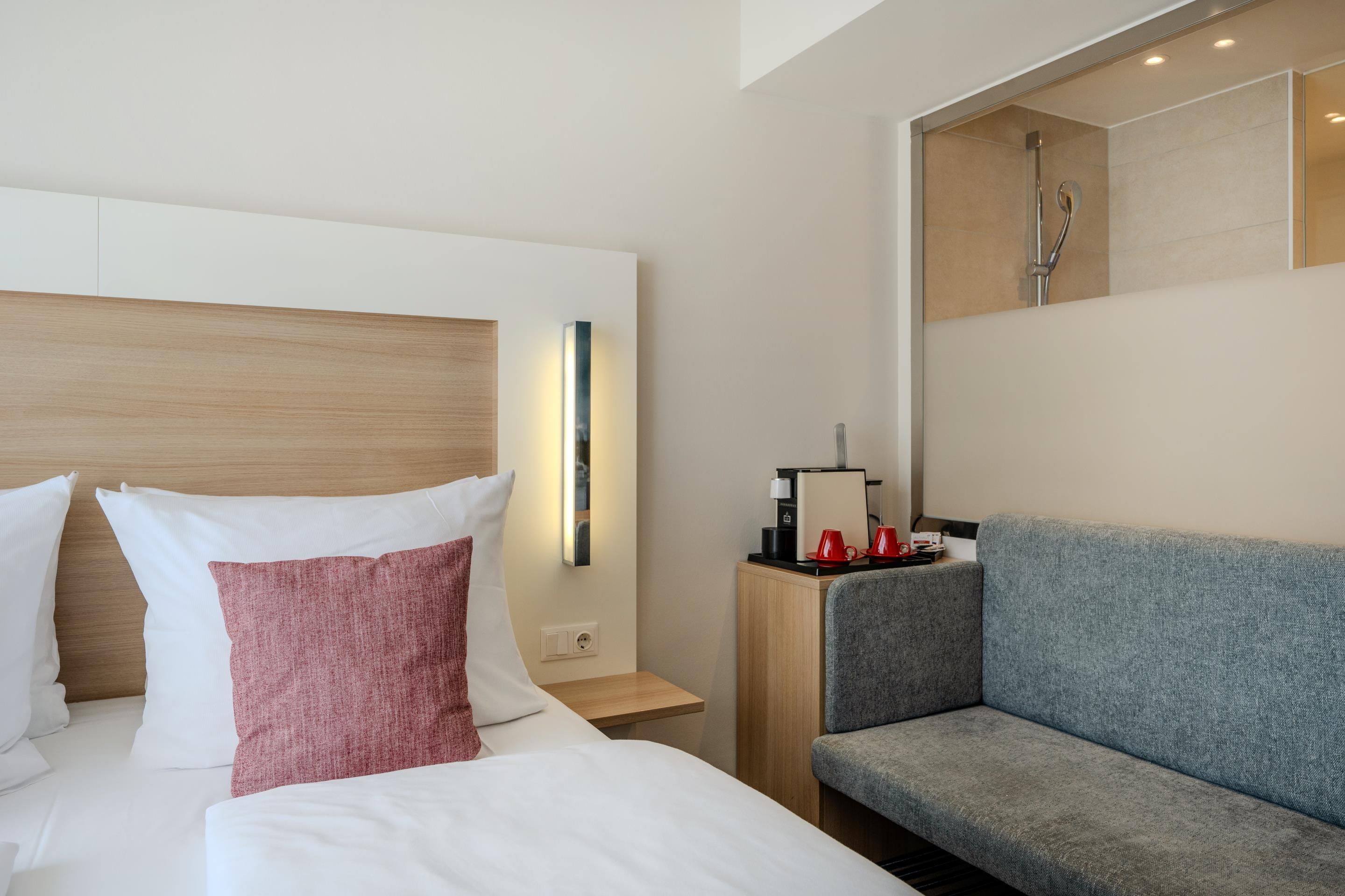 A modern hotel room with a bed, a grey sofa, a nightstand with a coffee maker, and a shower area.