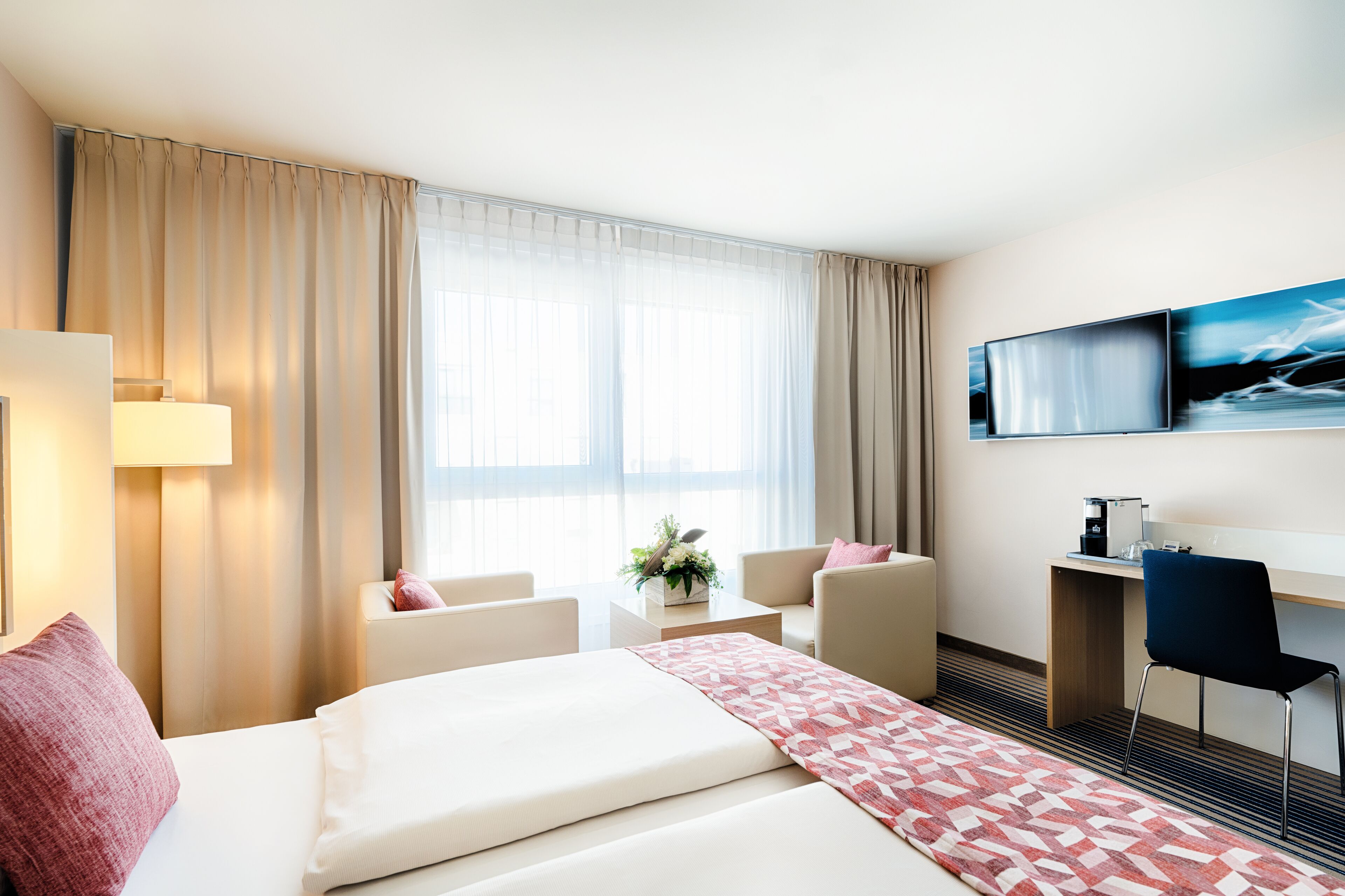 Una habitación de hotel con cama, escritorio, silla, televisor y ventana con cortinas.