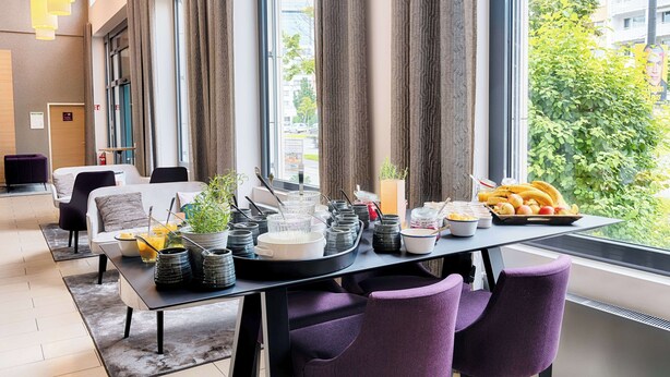 A modern dining area with a table set for breakfast, including fruits and beverages, near large windows with a city view.
