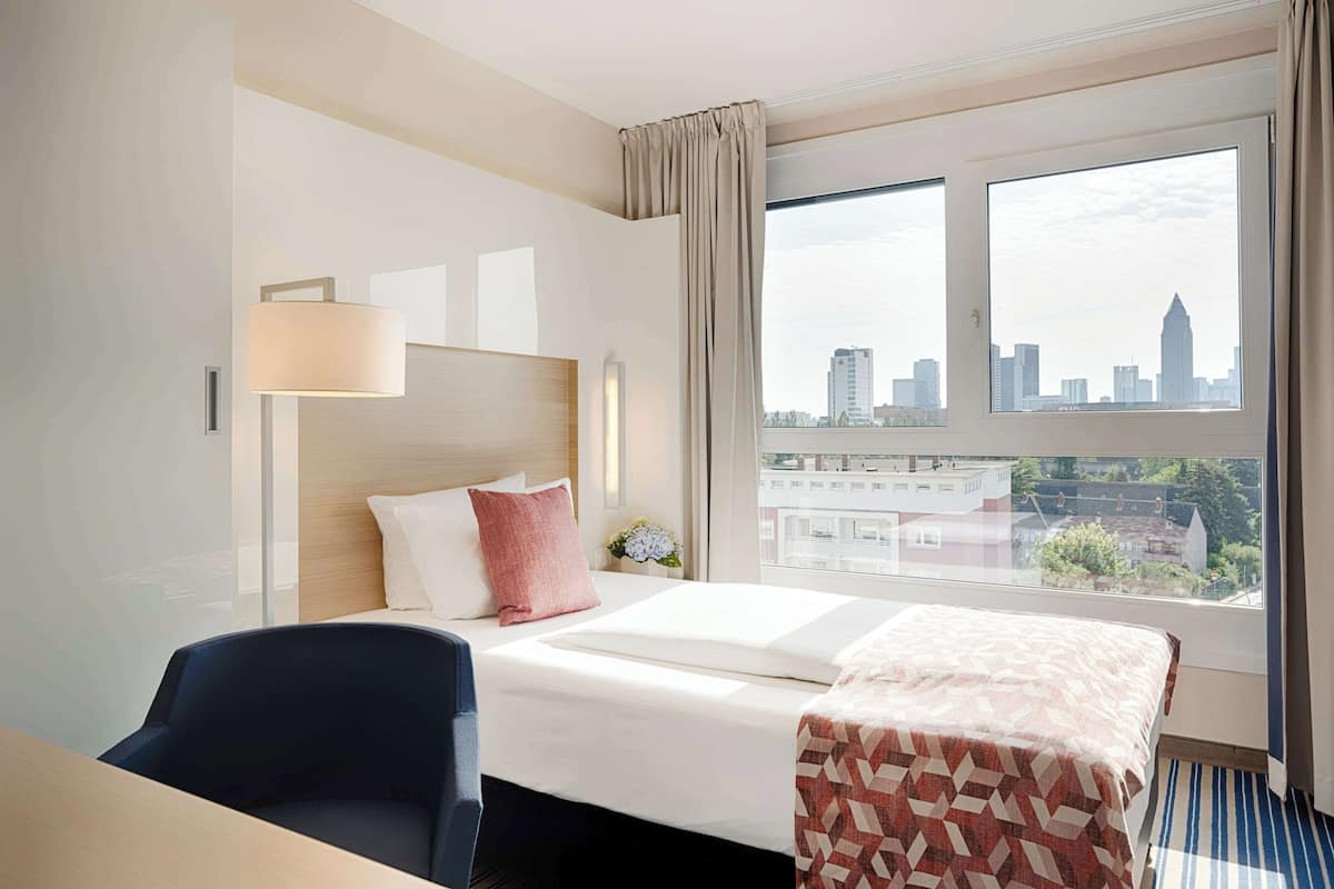 A hotel room with a bed, a desk, a chair, a lamp, and a large window with a city view.