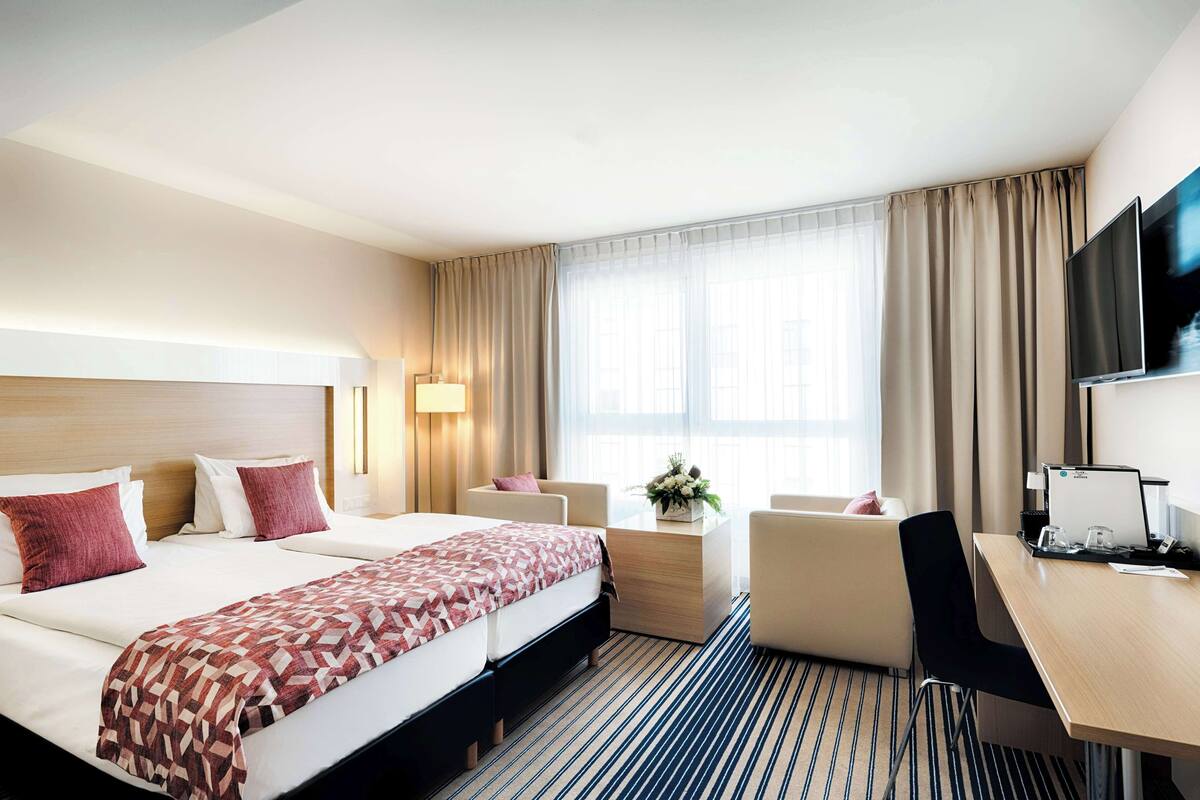 A hotel room with a large bed, a desk, a chair, a TV, and a window with curtains.