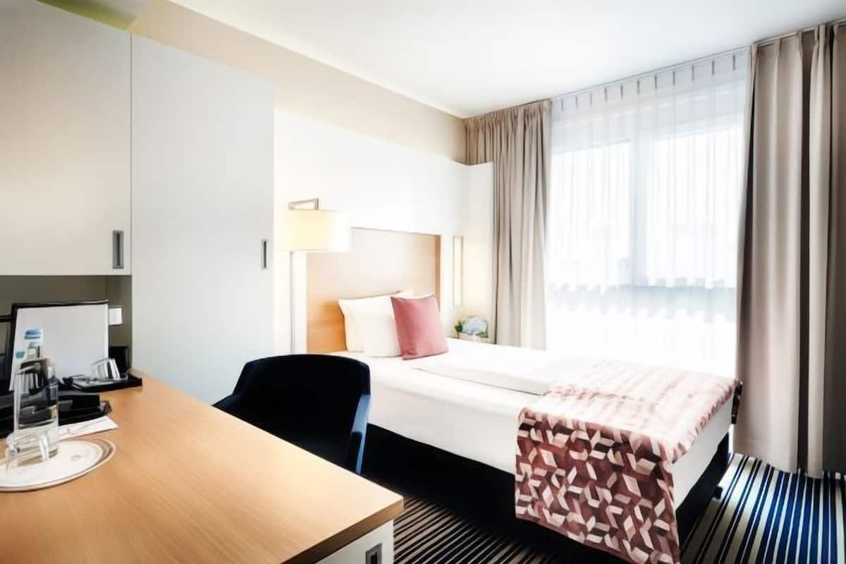 A hotel room with a bed, a desk with a computer, a chair, a lamp, and a window with curtains.