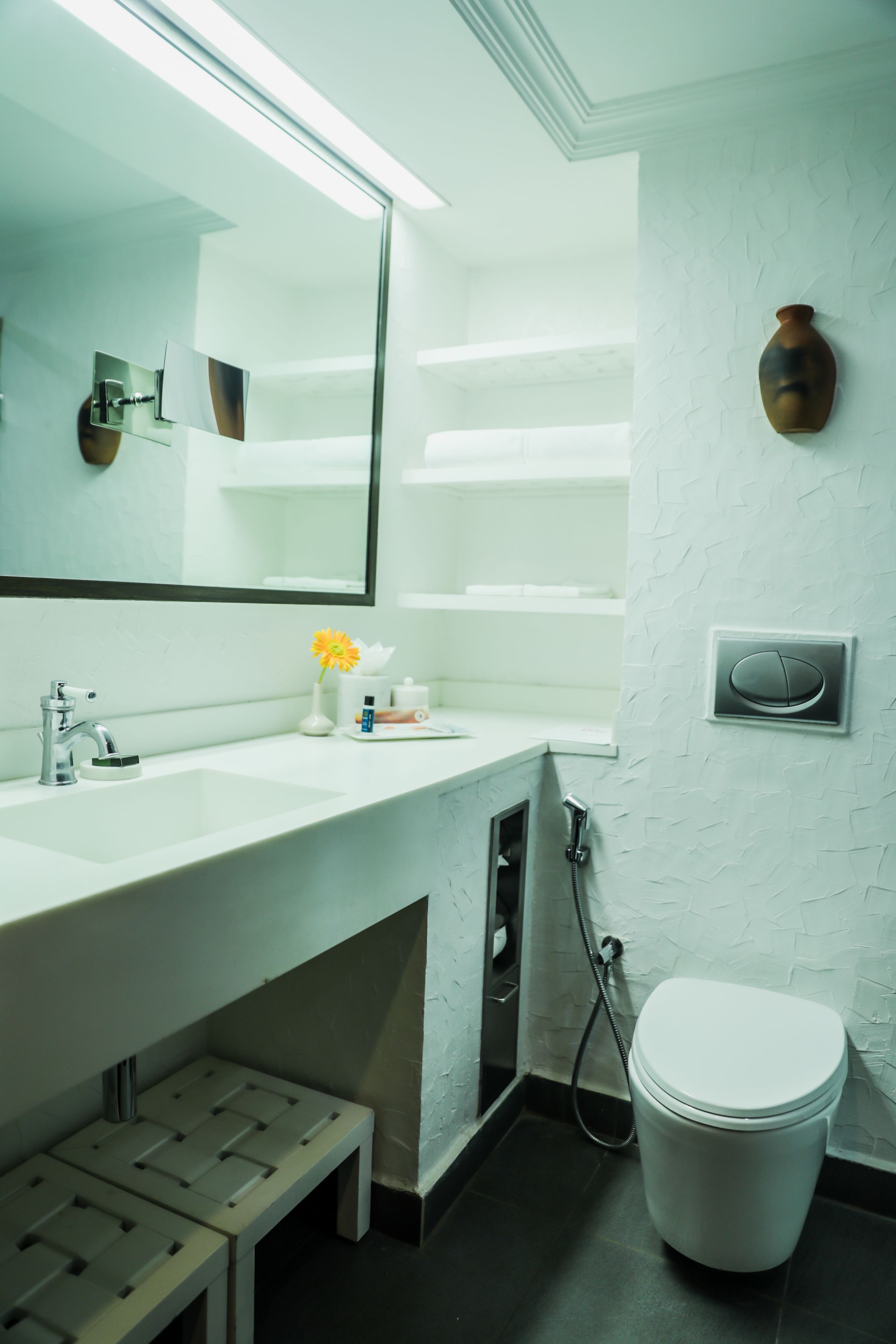 A bathroom with a white sink, mirror, and toilet. There is a small step stool under the sink and a showerhead next to the toilet.
