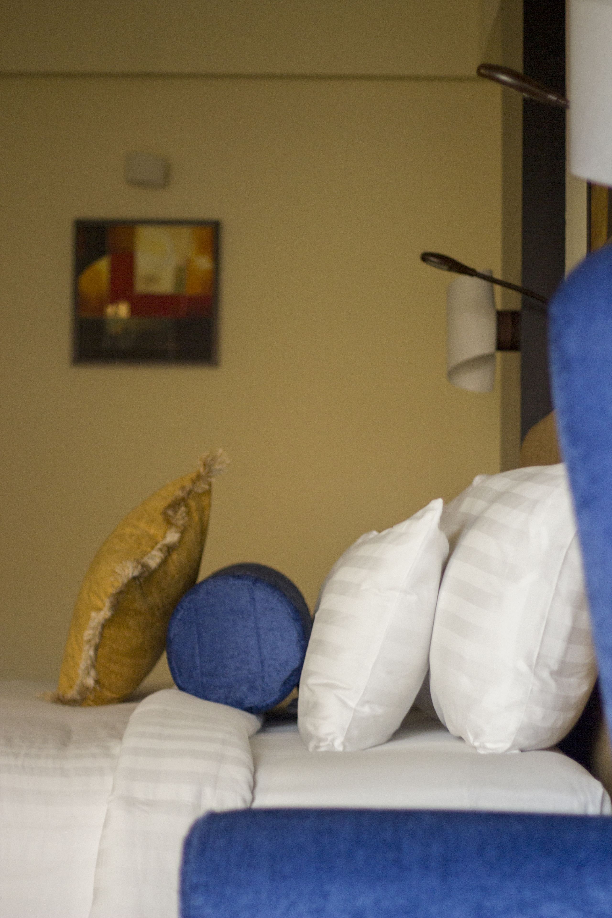 Una cama con almohadas blancas y azules, una pared color crema y un cuadro enmarcado.