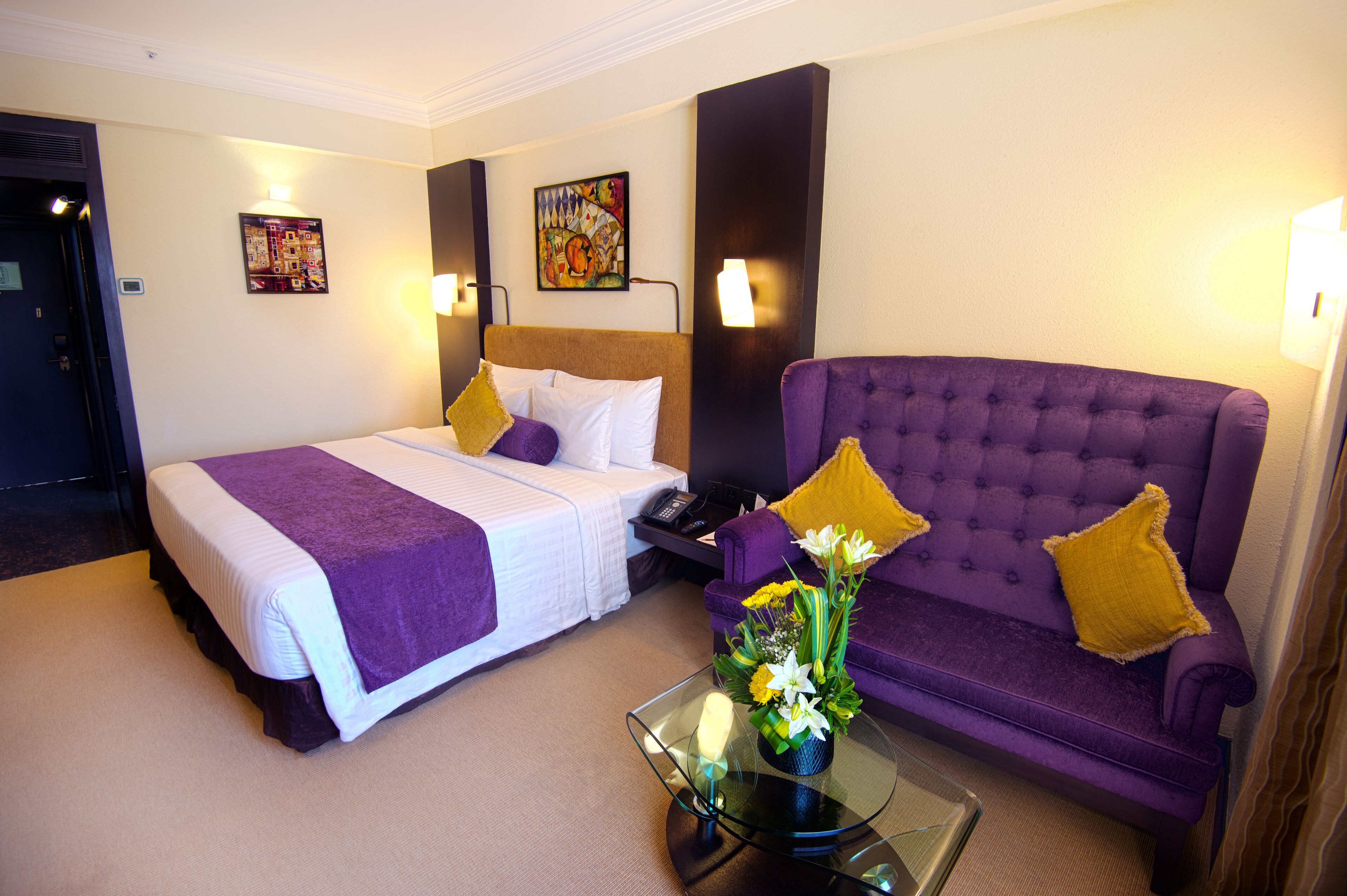 A hotel room with a bed, a purple sofa, and a glass coffee table with a flower arrangement.