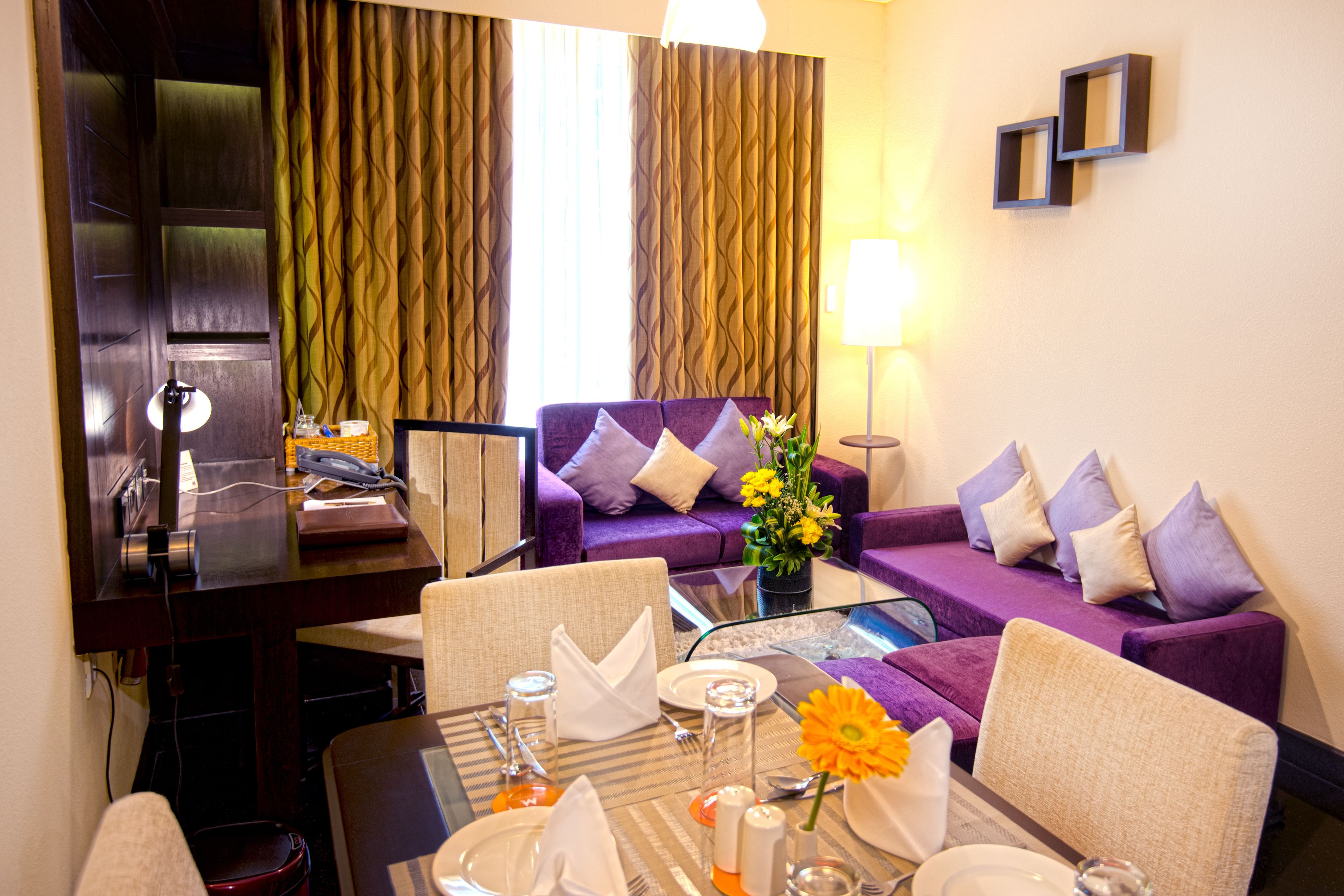A hotel room with a dining area, a purple sofa, a desk with a lamp, and a view of the outside through curtains.