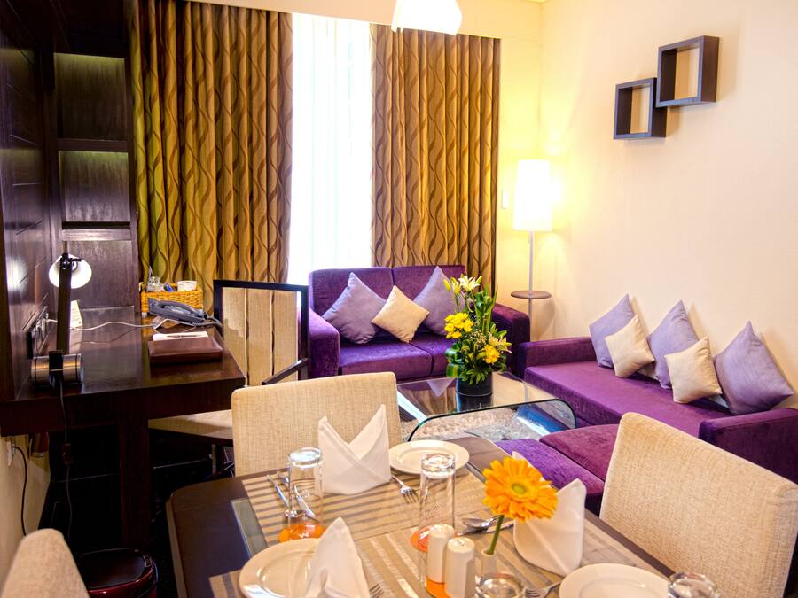 A hotel room with a dining area, a purple sofa, a desk with a lamp, and a view of the outside through curtains.