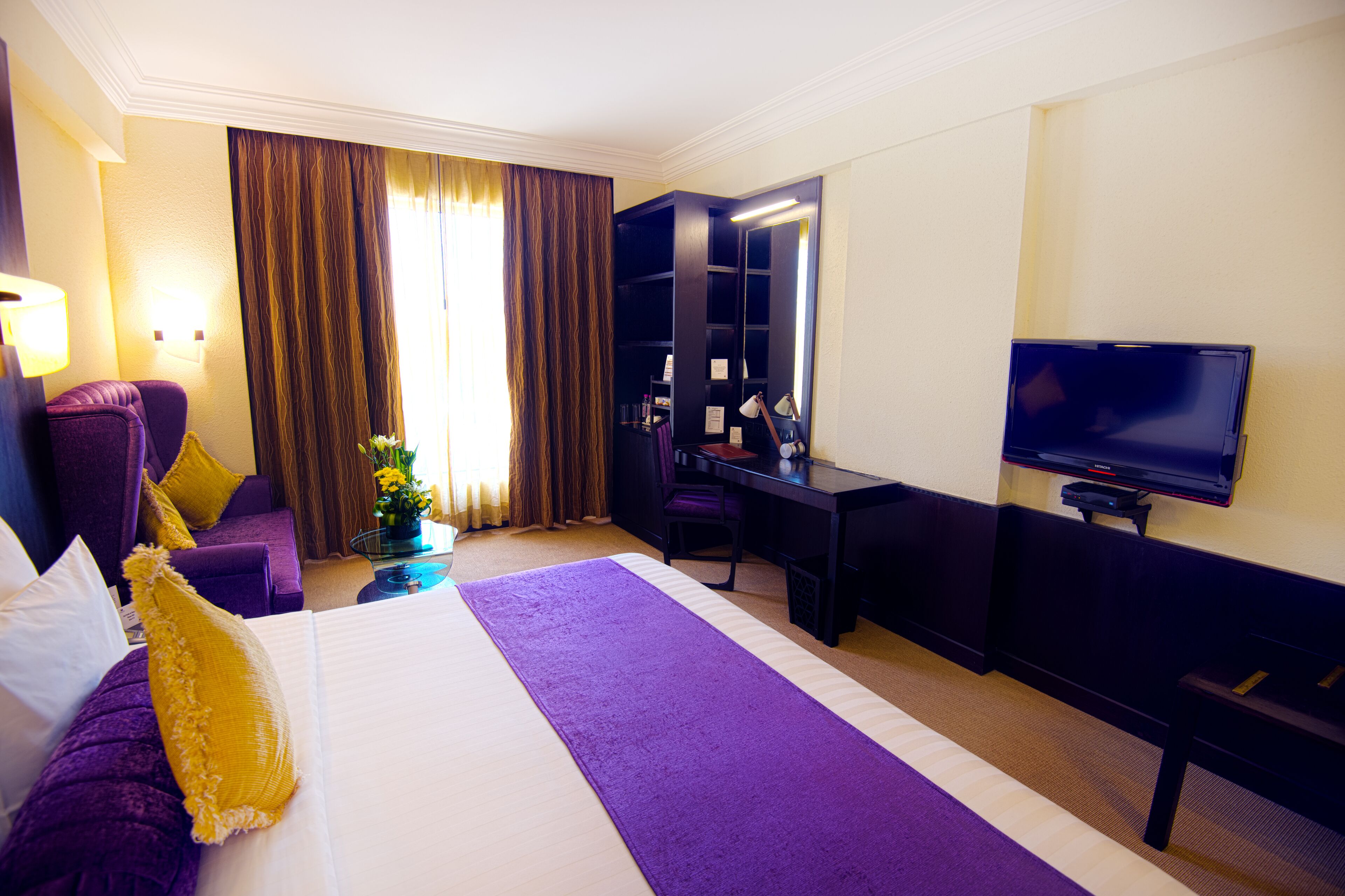 A hotel room with a large bed, a desk, a television, and a seating area with purple furniture.