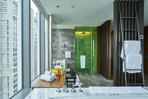 A modern bathroom with a large mirror, a sink, a shower, and a toilet. There is a window providing a view of the city.