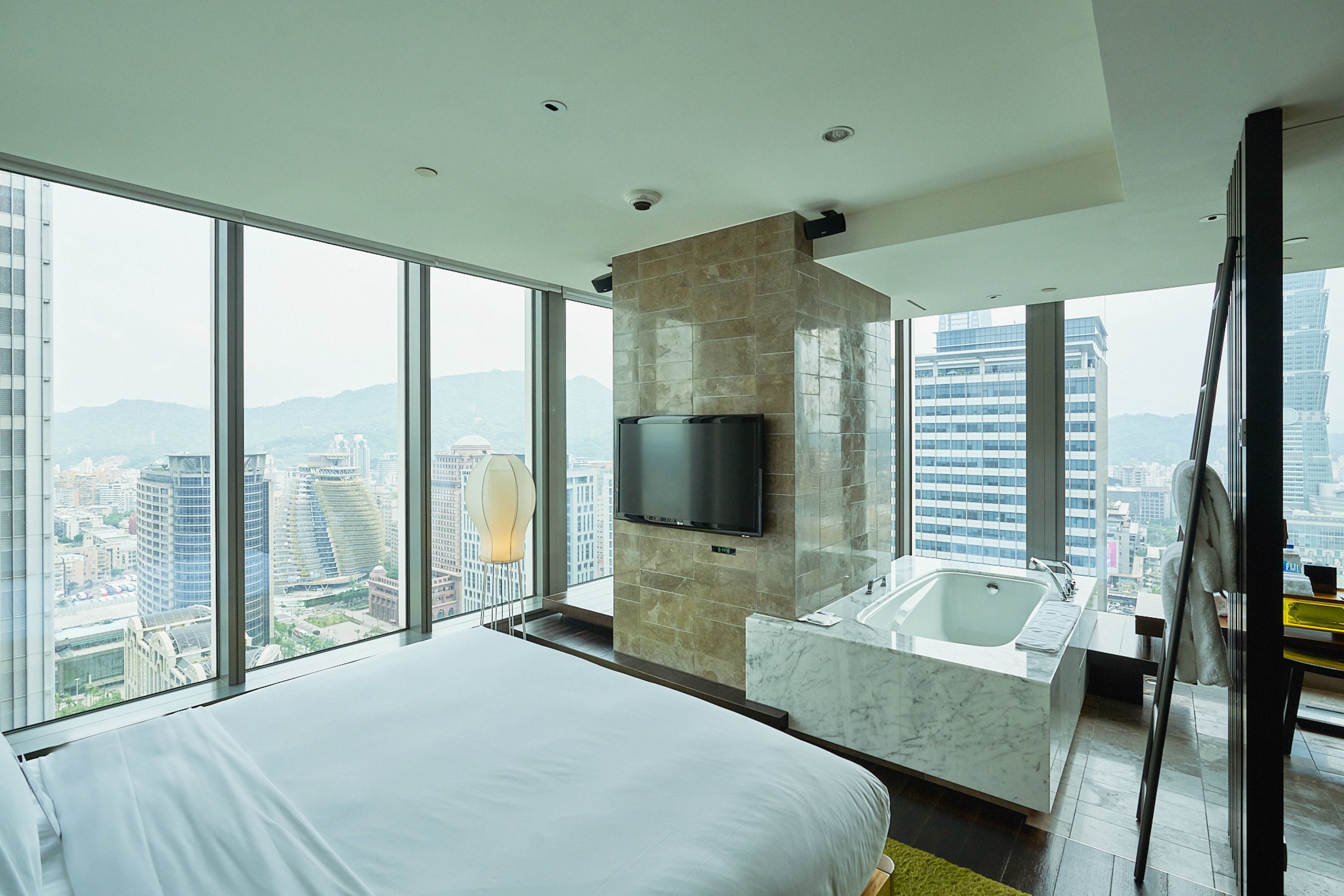 A modern hotel room with a large bed, a built-in TV, a marble bathroom, and a city view.