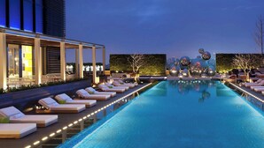 Zona de piscina moderna de hotel con sillones de descanso y vista a un paisaje urbano al atardecer.