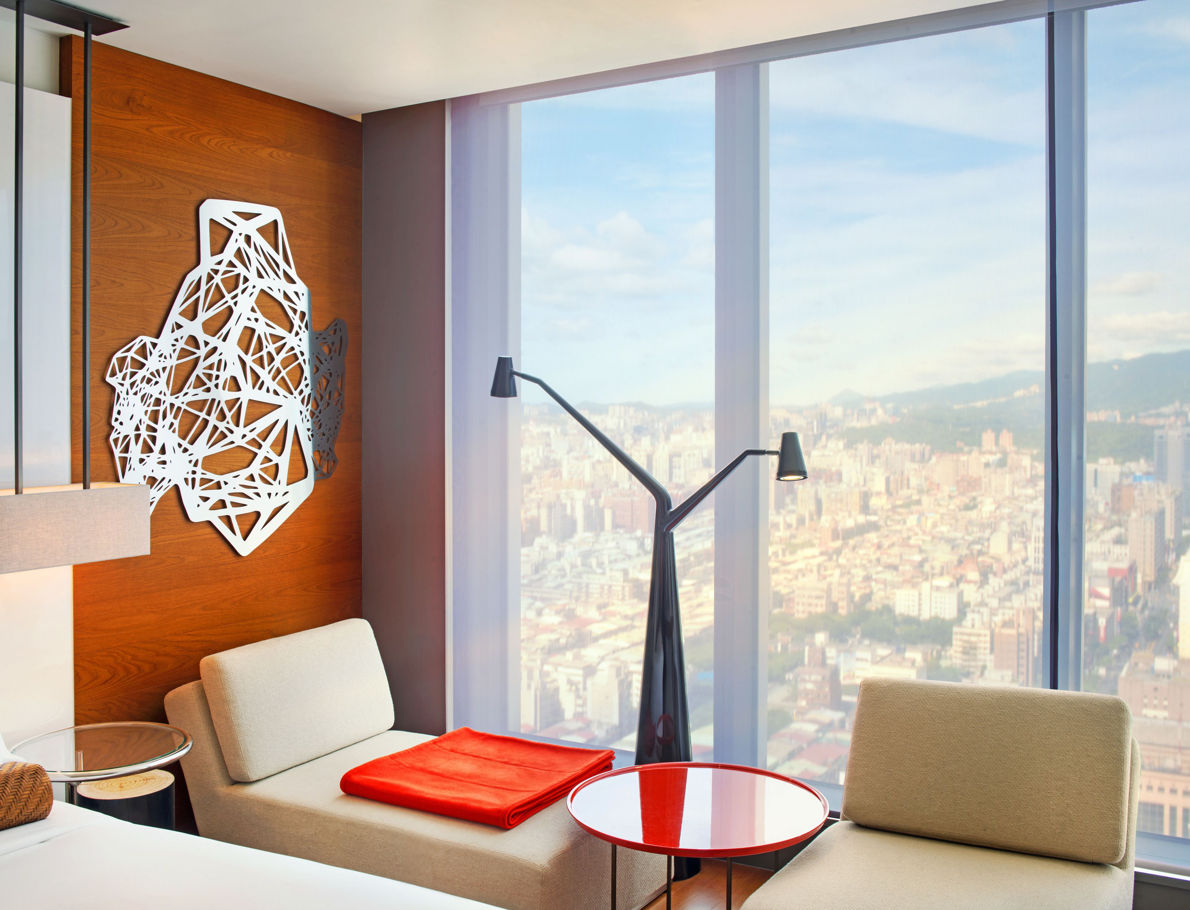 A modern hotel room with a large window offering a city view, a sofa with a red throw, and a unique wall art piece.
