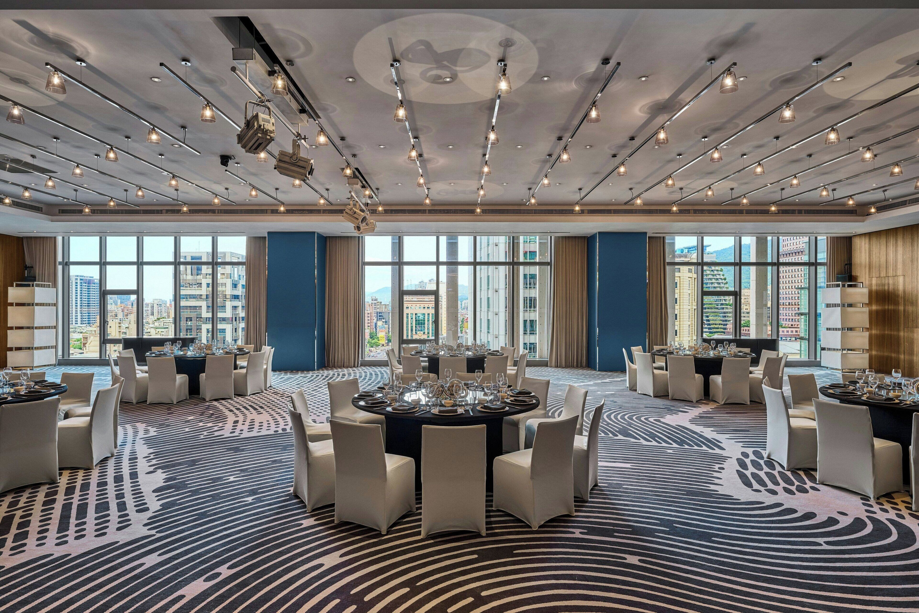 A spacious conference room with a modern ceiling design, large windows offering a city view, and a patterned carpet.