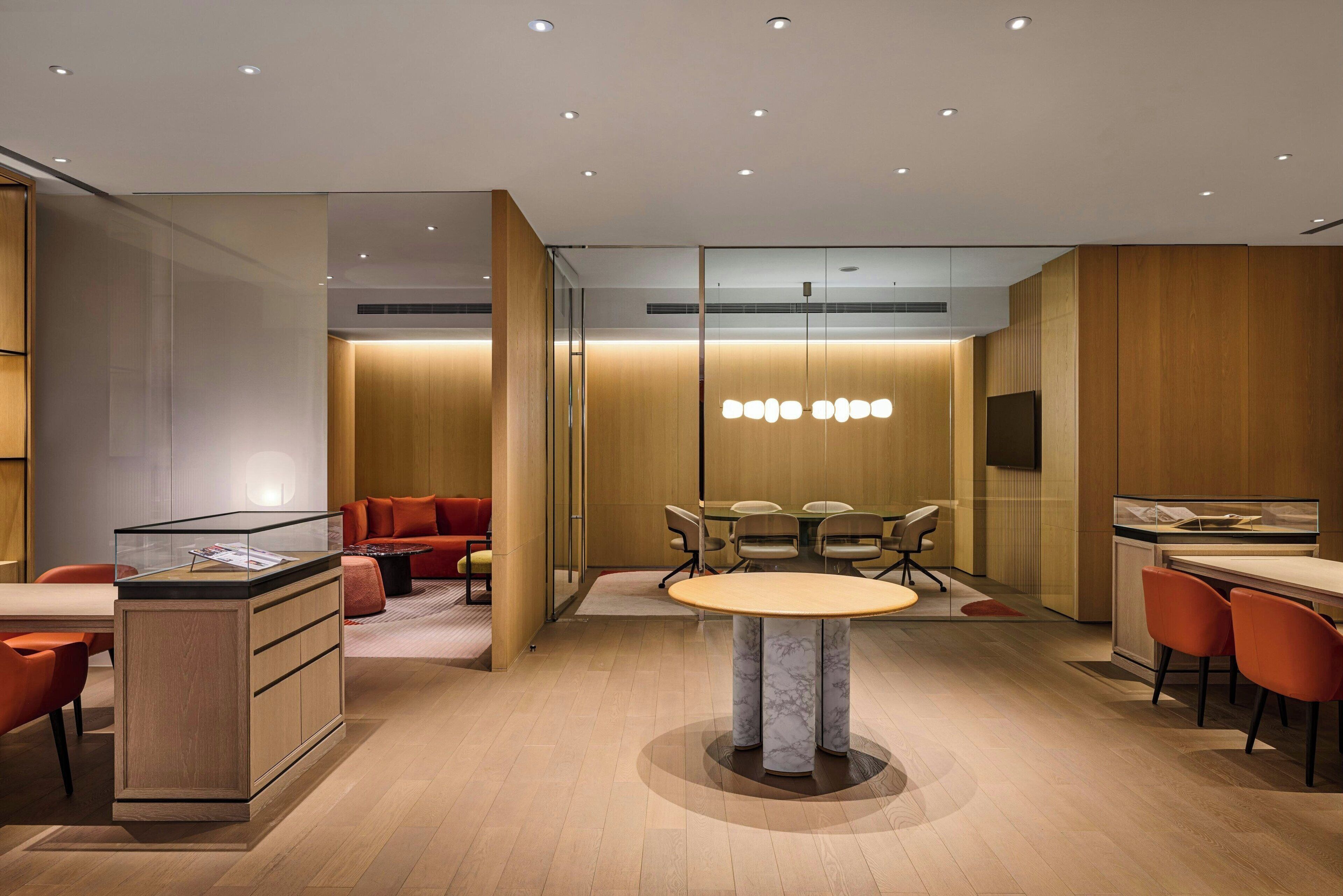 A modern office space with a glass partition, a round table with chairs, and a seating area with red chairs.