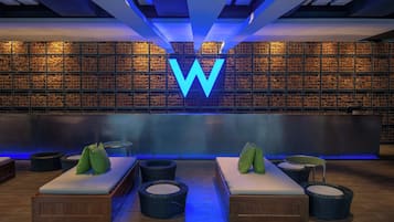 A modern lounge area with a large illuminated 'W' logo, wooden benches, and round wicker chairs.