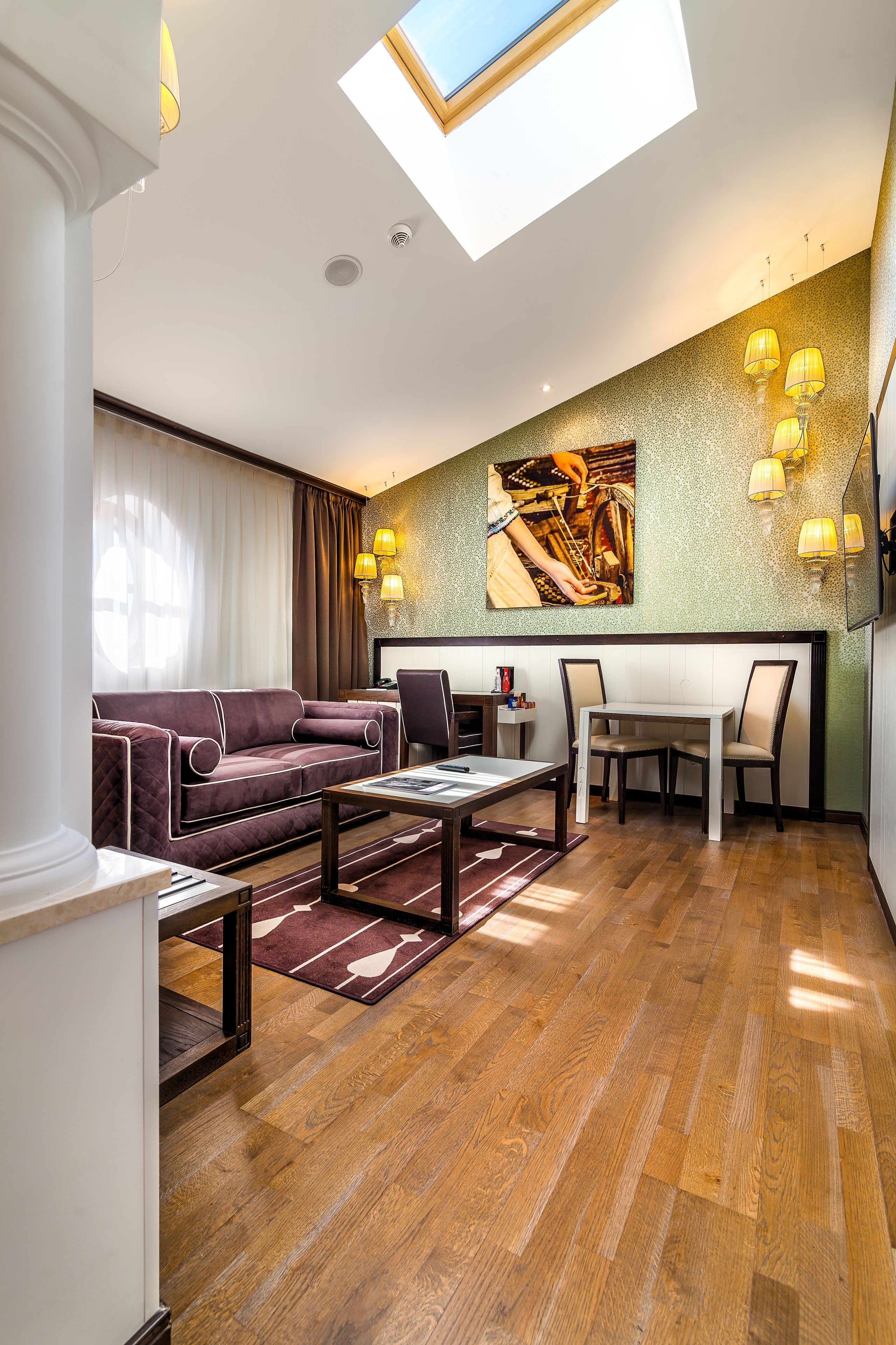 A modern living room with a sofa, dining table, and chairs. There is a skylight above the dining area and a painting on the wall.