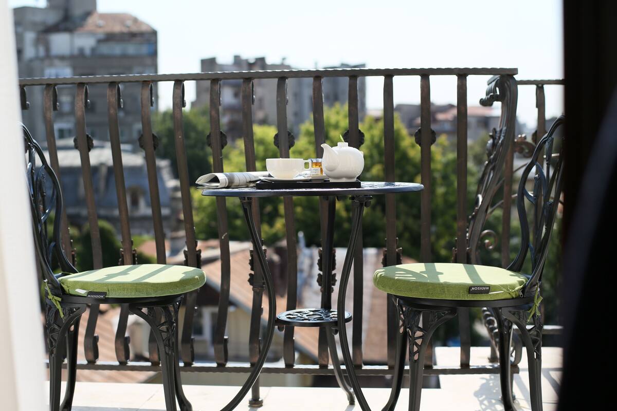 A balcony with a table set for tea, two chairs, and a view of buildings and trees.