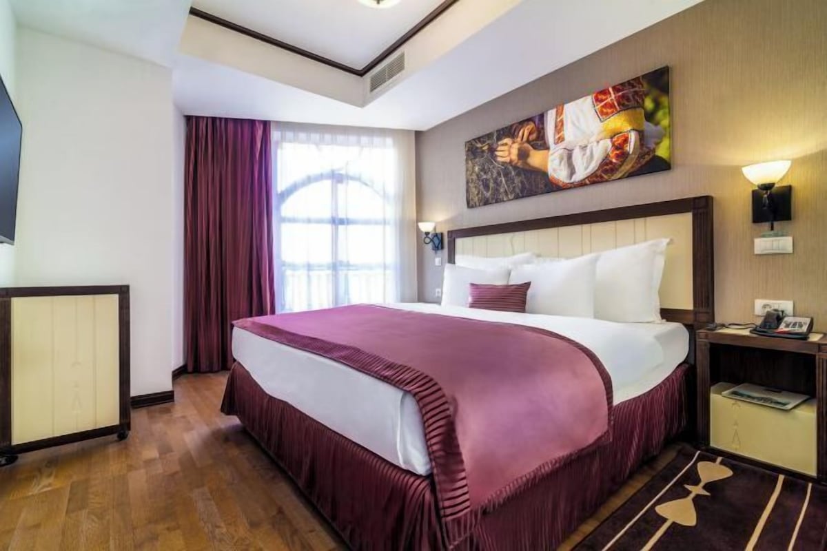 A hotel room with a large bed, a painting on the wall, a nightstand, a lamp, and a window with curtains.