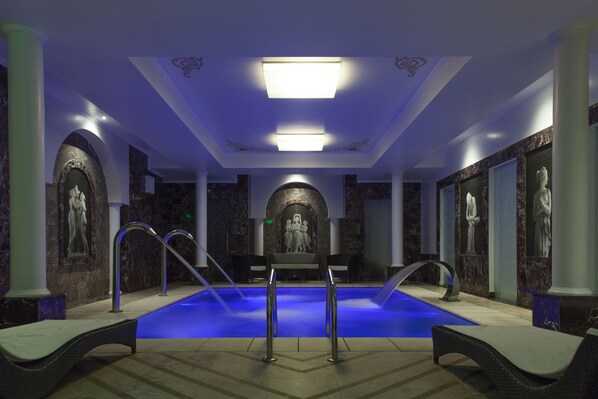 A modern indoor pool area with a blue-lit pool, marble walls, and classical statues.
