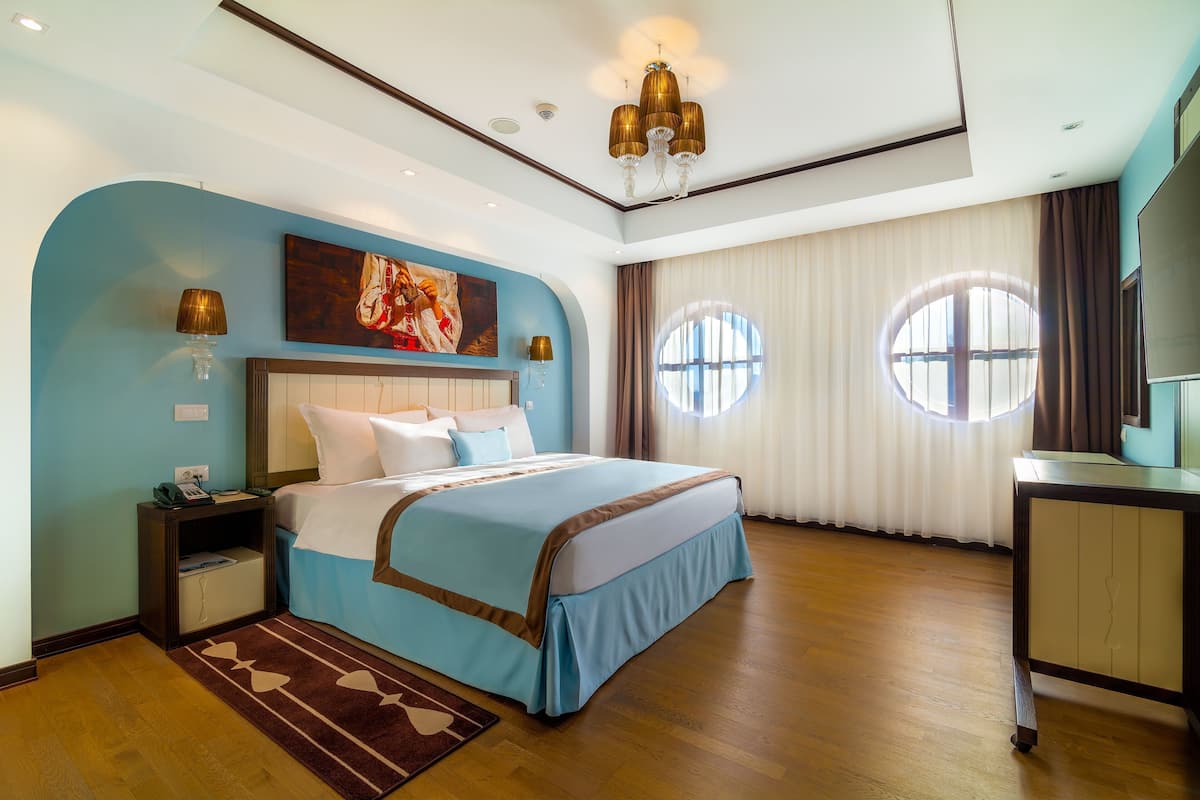 A bedroom with a bed, bedside tables, a nightstand, a desk, and a large window.