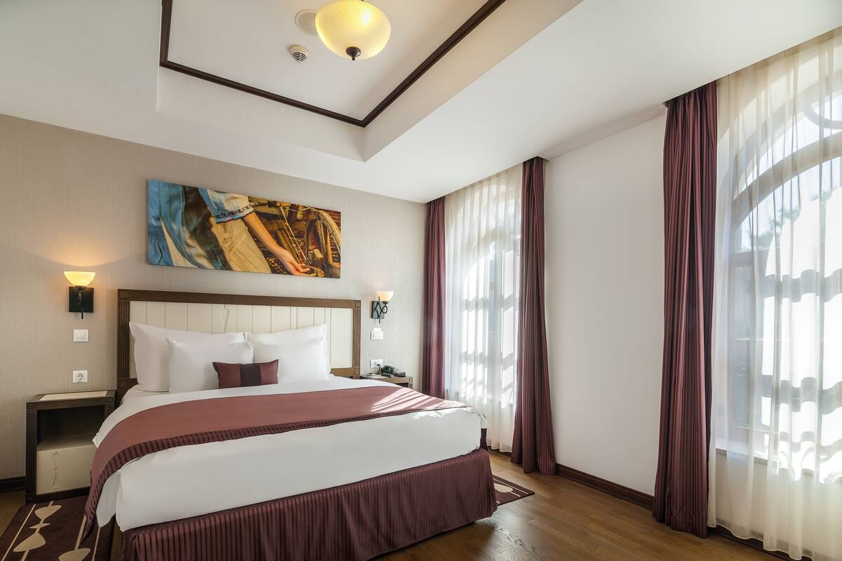 A hotel room with a large bed, a headboard, a painting, a nightstand, a lamp, and sheer curtains.