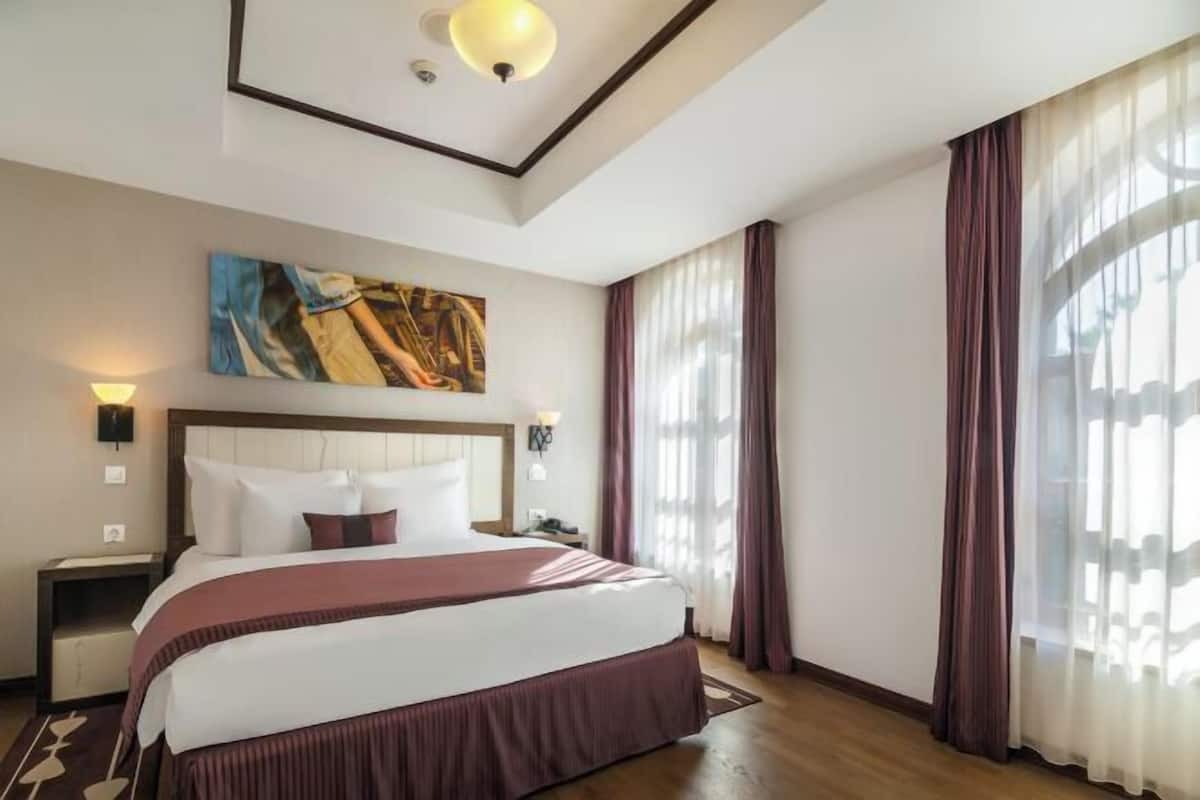 A hotel room with a large bed, a headboard, a painting on the wall, and a window with curtains.