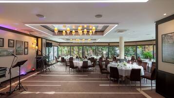 A spacious dining area with multiple tables set for guests, large windows offering a view of the outdoors, and a stage with musical equipment.