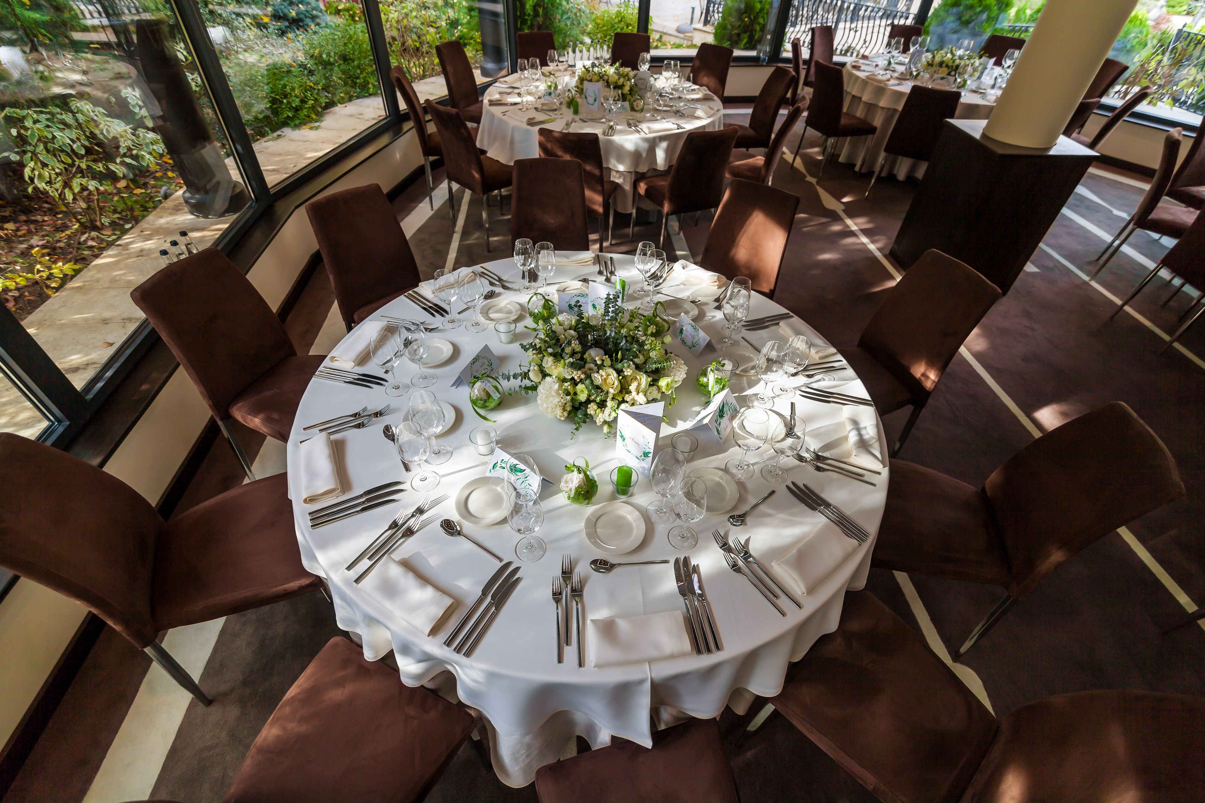 A round dining table set for six with white tablecloths, brown chairs, and a floral centerpiece.