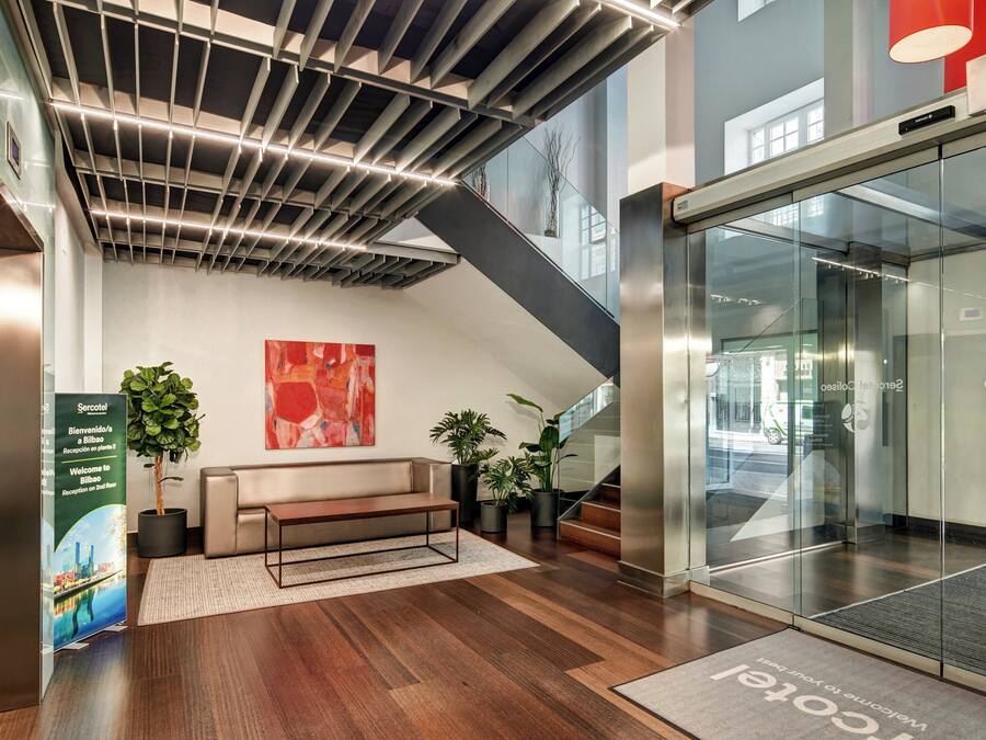 A modern reception area with a bench, potted plants, a glass elevator, and a welcome mat.