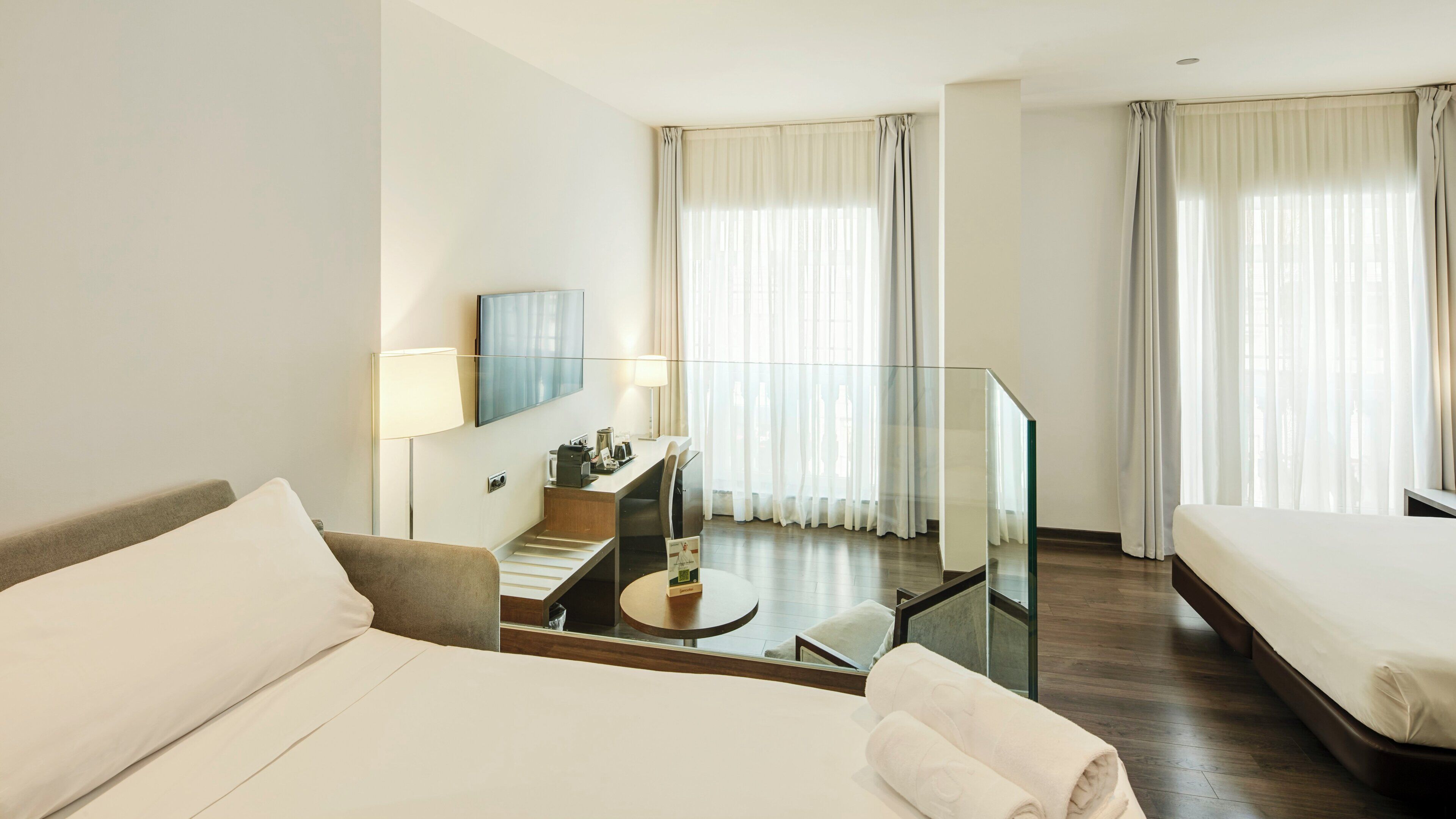 A hotel room with a bed, a desk with a lamp, a chair, a TV, and a glass railing.
