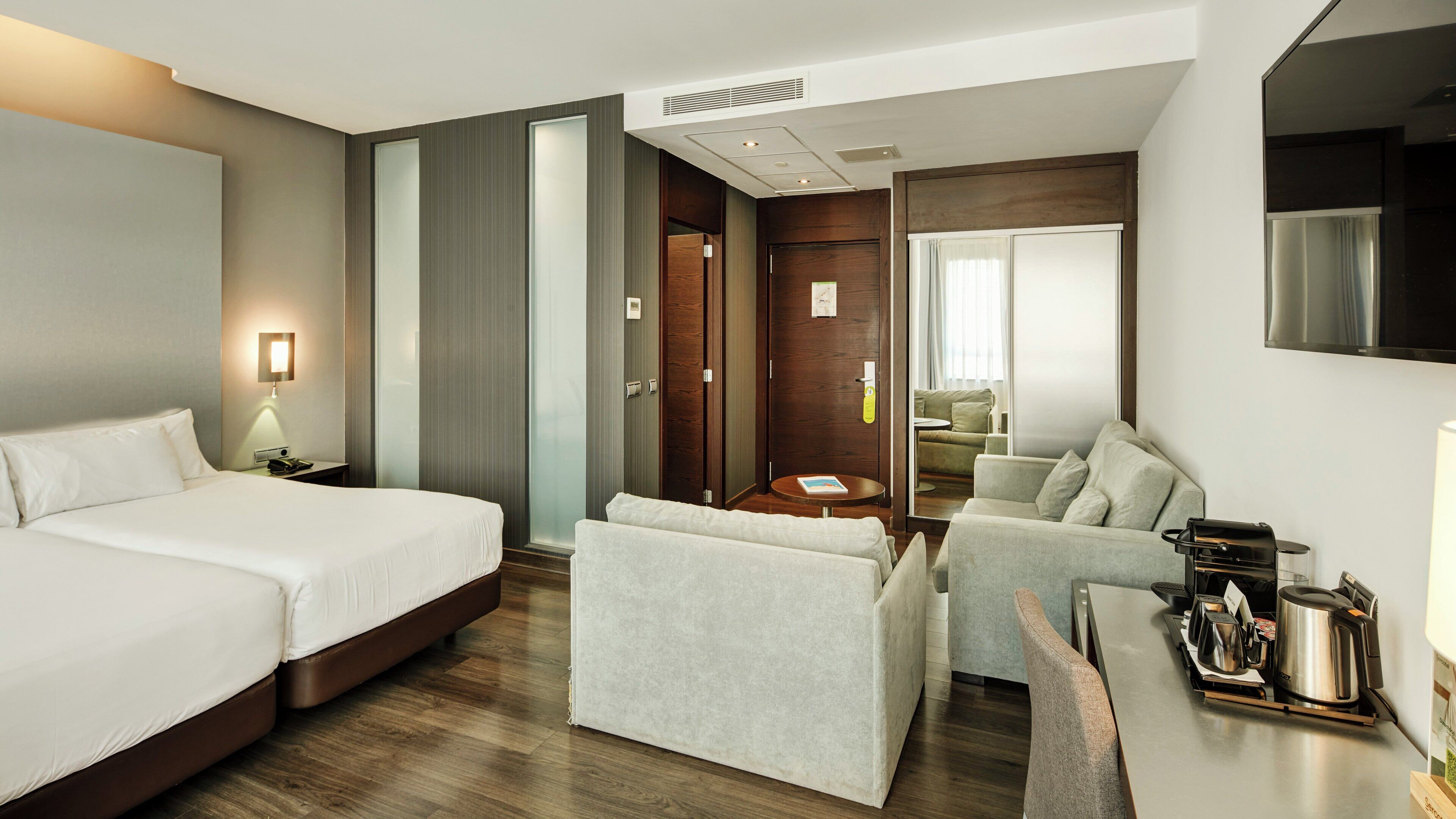 A hotel room with a bed, a desk with a coffee maker, a sofa, and a view of the city.