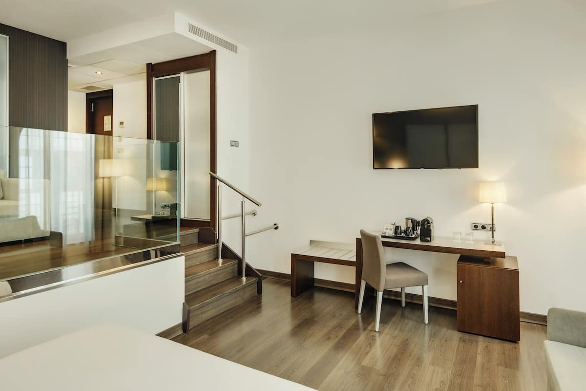 Ein modernes Hotelzimmer mit einer Holztreppe, einem Flachbildfernseher, einem Schreibtisch mit Stuhl, einem Sofa und einer Lampe.