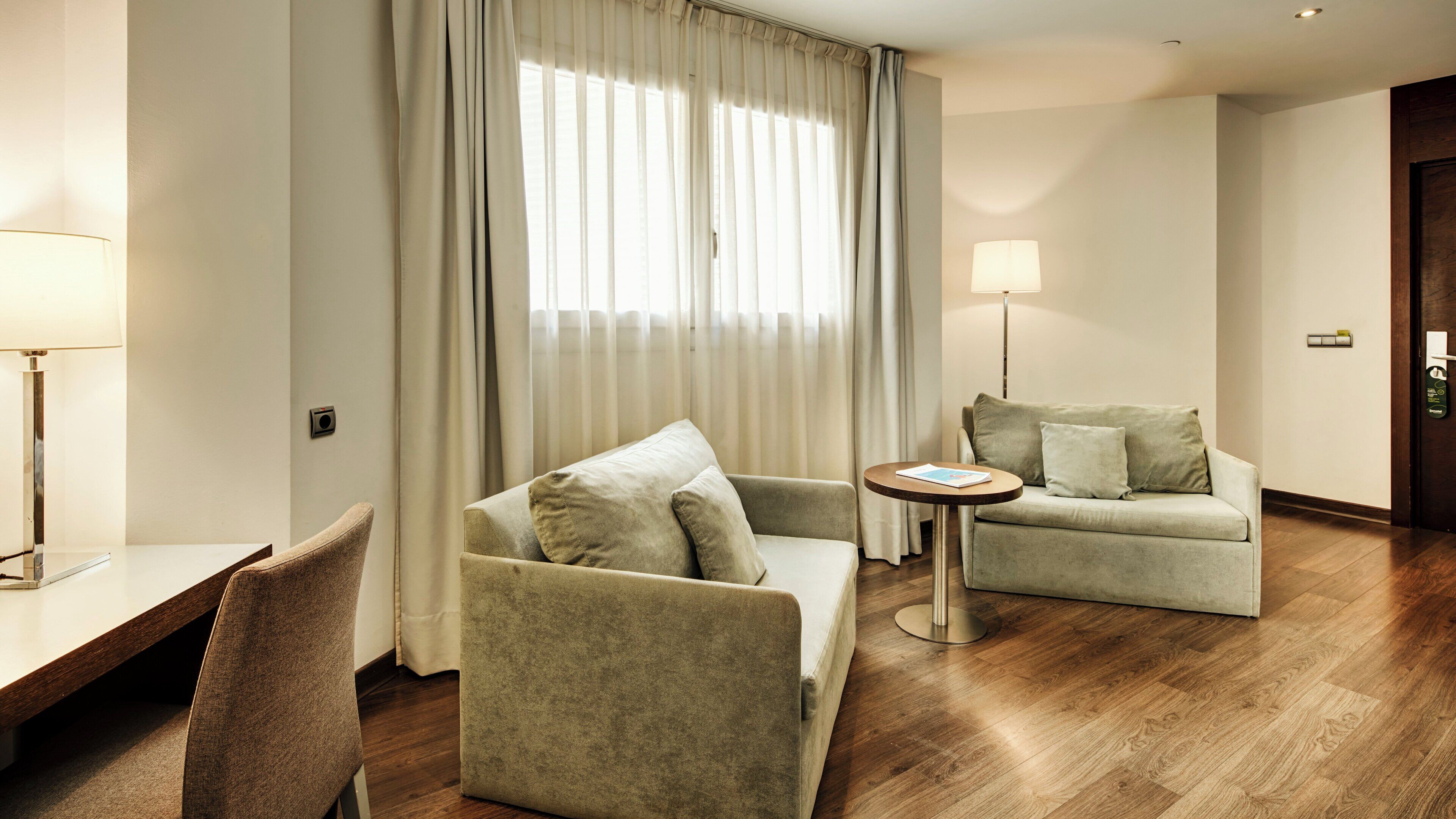 A hotel room with a sofa, armchair, small round table, and a lamp.